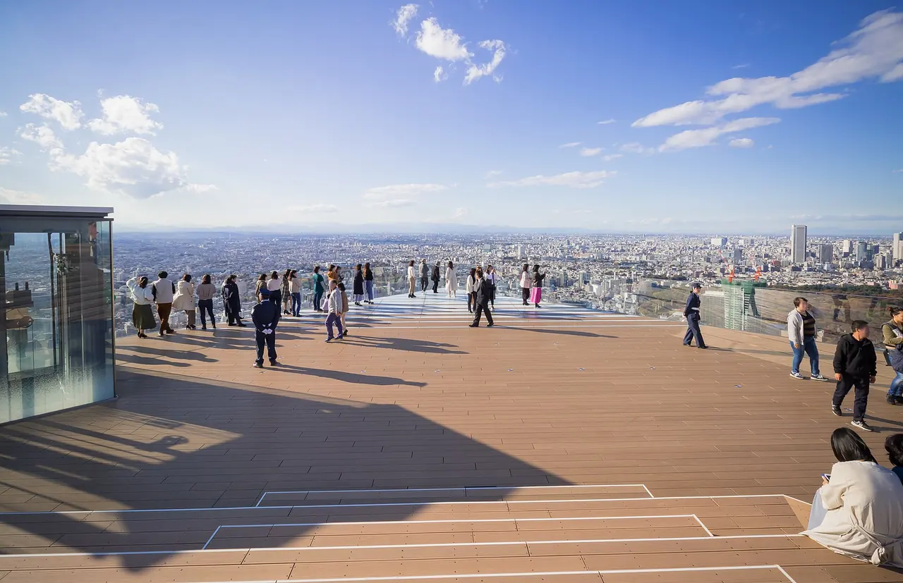 Narożnik Sky Edge na dachu SHIBUYA SKY w pogodne popołudnie, z gośćmi przy szklanej barierze i Tokio rozciągającym się aż po horyzont