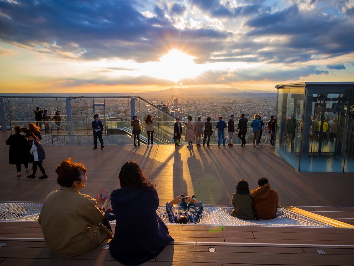Otwarty dach SHIBUYA SKY o zachodzie słońca, z ludźmi na tarasie i szklaną krawędzią, oraz słońcem nisko nad panoramą Tokio